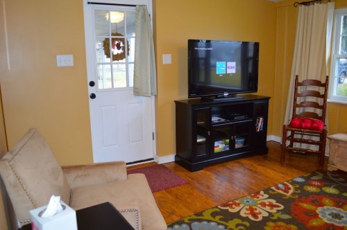 Cozy living room with a TV, chair, and door, featuring warm yellow walls and a colorful rug.