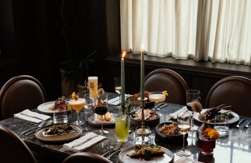 A beautifully set dining table with various gourmet dishes, drinks, and candles, illuminated by soft natural light.