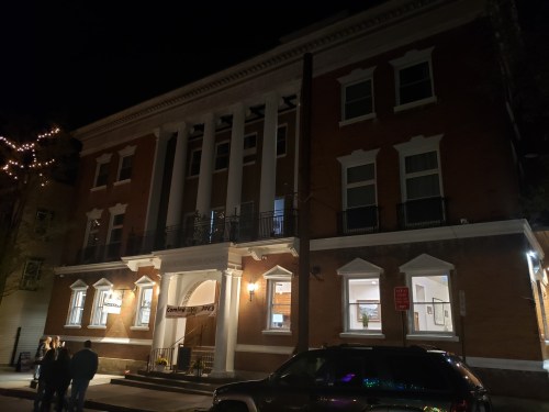 A historic brick building with columns, illuminated at night, with people gathered outside and a parked car in front.