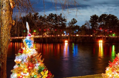 A decorated Christmas tree by a river, reflecting colorful lights from the opposite bank under a twilight sky.