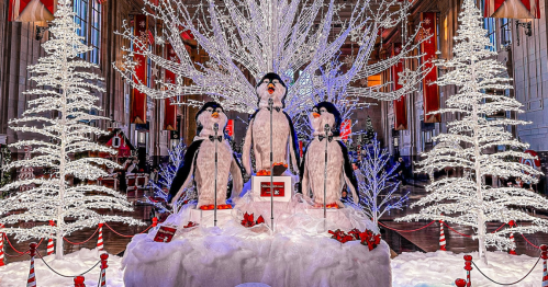 Three festive penguin figures stand on a snowy display, surrounded by sparkling white trees and holiday decorations.