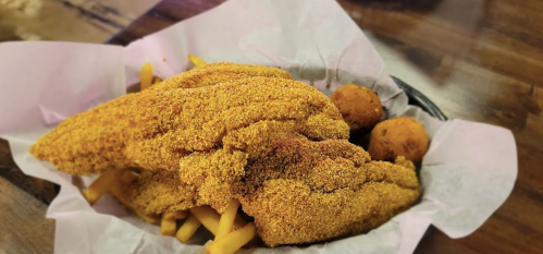 A basket of golden fried fish fillets on a bed of fries, accompanied by two hushpuppies.