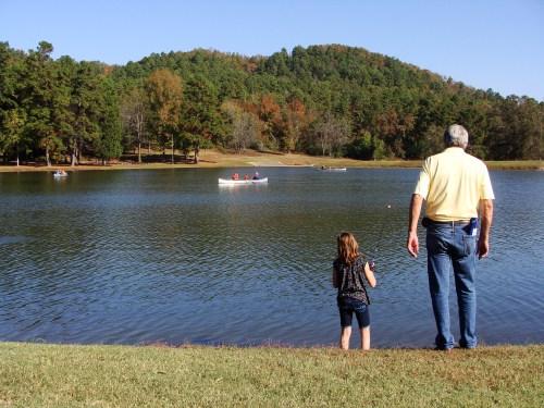 A man and a girl stand by a lake, watching boats on the water with trees and a hill in the background.