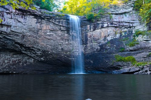 A serene waterfall cascading over rocky cliffs into a calm pool, surrounded by lush greenery and autumn foliage.