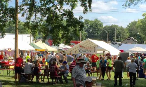A lively outdoor festival scene with tents, food vendors, and crowds enjoying the event under a sunny sky.