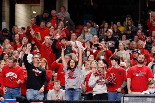 A lively crowd of fans in red and black cheer enthusiastically at a sporting event, waving pom-poms and celebrating.