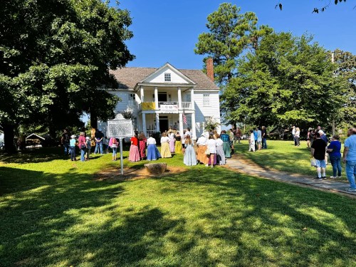 A historic house with people in period costumes gathered on the lawn, enjoying a sunny day.