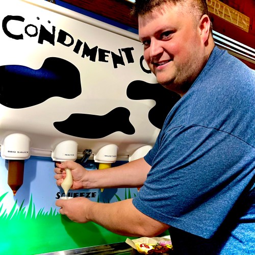 A man smiles while using a condiment dispenser shaped like a cow, squeezing out mayo.