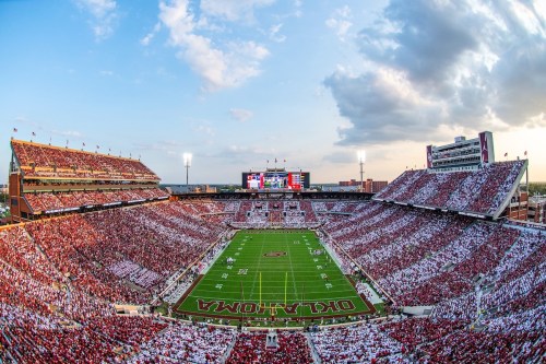 A panoramic view of a packed football stadium at sunset, filled with fans in red and white, with a scoreboard visible.