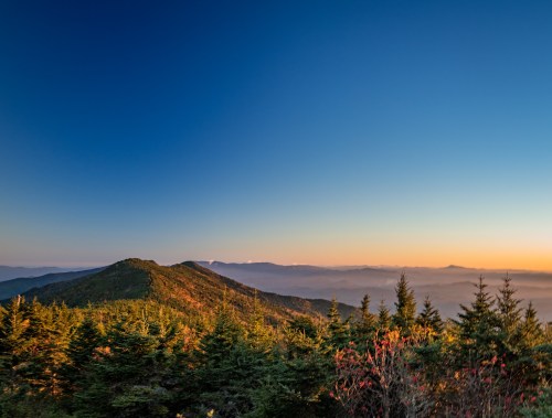 A panoramic view of mountains under a clear blue sky, with lush green trees in the foreground and a warm sunset glow.