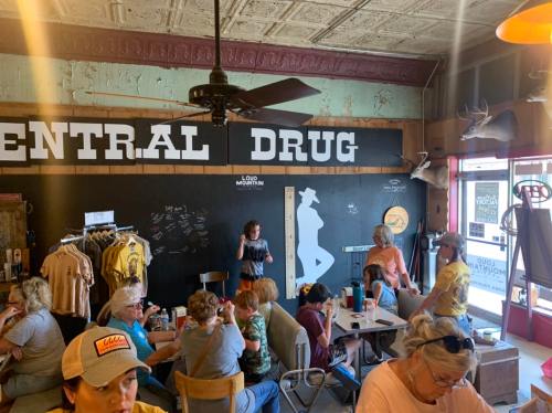 A busy café interior with a "Central Drug" sign, people chatting, and merchandise displayed on the walls.