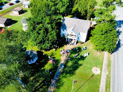 Aerial view of a historic house surrounded by trees, with a gathering of people on the lawn and nearby buildings.
