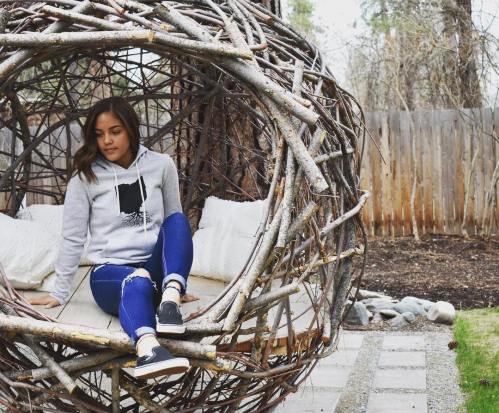 A young woman sits in a large, woven nest-like chair in a garden, wearing a hoodie and jeans.