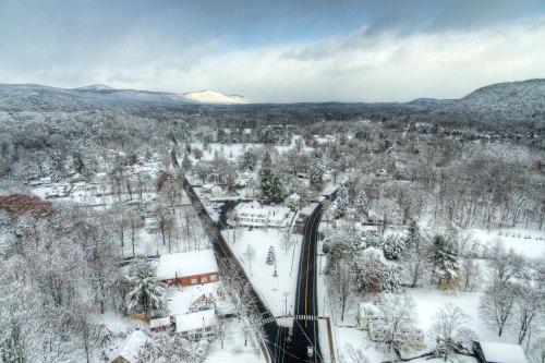 A snowy landscape with a winding road, surrounded by trees and houses, under a cloudy sky. Mountains in the distance.