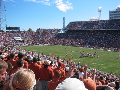 A packed stadium with fans in orange cheering as a football game unfolds on a sunny day.