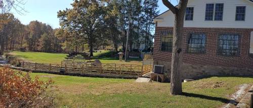 A peaceful outdoor scene featuring a grassy area, wooden bridge, and a house surrounded by trees in autumn.