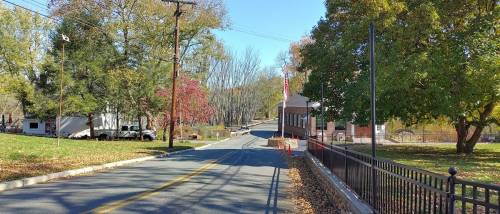 A quiet road lined with trees, leading to a bridge and a building, with autumn leaves scattered on the ground.