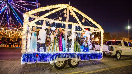 A festive parade float decorated with lights, featuring performers in colorful costumes and a winter theme.
