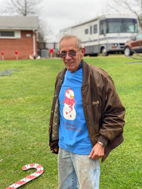 An older man wearing sunglasses and a festive blue shirt stands on a lawn, smiling in front of a house and RV.