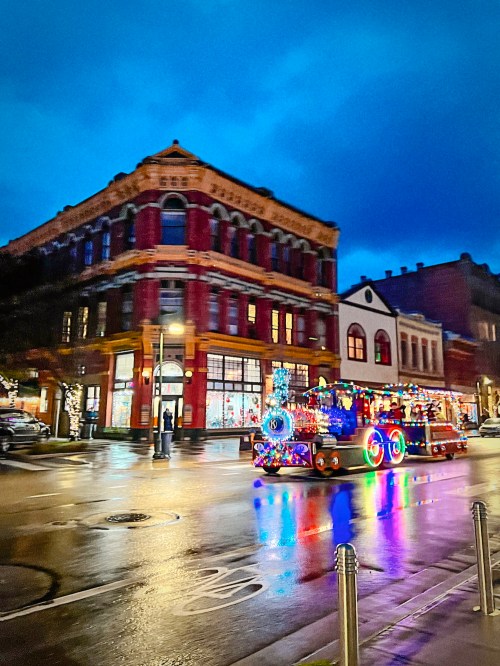 A brightly lit, decorated vehicle drives past a historic building at dusk, reflecting colorful lights on wet pavement.