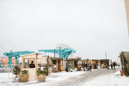 A snowy street scene with colorful market stalls and a blue bridge in the background. People are walking around.