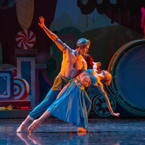 A male and female ballet dancer perform a dramatic lift on stage, surrounded by colorful set designs.