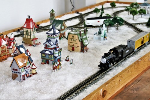A festive miniature train set with colorful buildings and snowy scenery, featuring a black steam engine on the tracks.