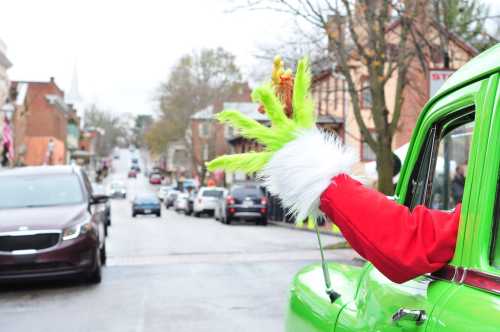 A green hand in a red sleeve waves from a car, with a festive street scene in the background.