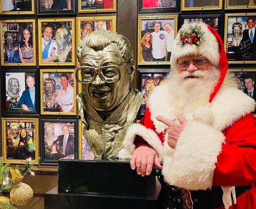 Santa Claus poses next to a bust in a gallery filled with framed photos of various people.
