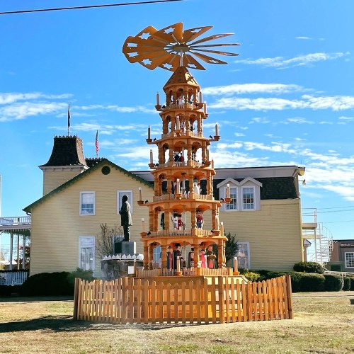 A tall wooden Christmas pyramid with multiple tiers, surrounded by a fence, set against a clear blue sky and buildings.