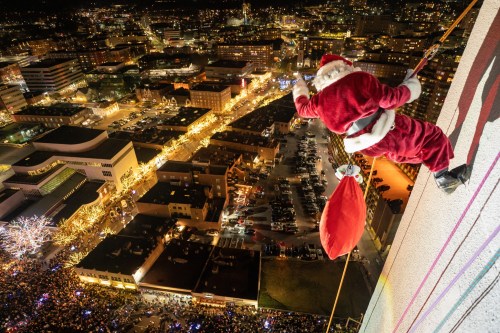 Santa Claus rappelling down a tall building, overlooking a festive cityscape filled with lights and a crowd below.