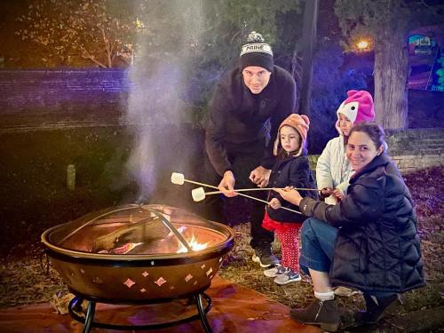 A family gathers around a fire pit, roasting marshmallows on sticks, with festive lights in the background.