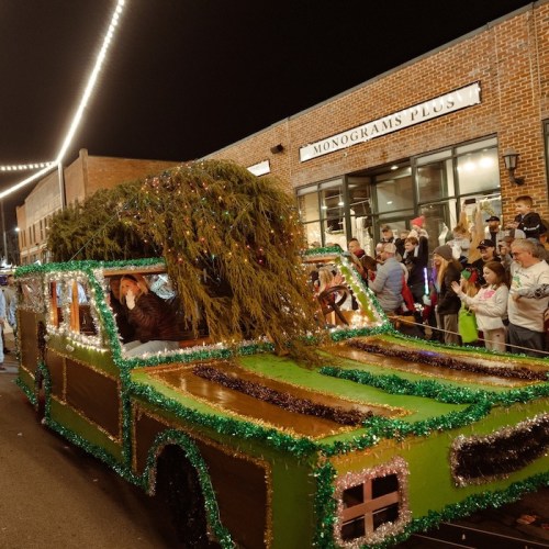 A festive parade float decorated with greenery and lights, surrounded by spectators on a lively street at night.