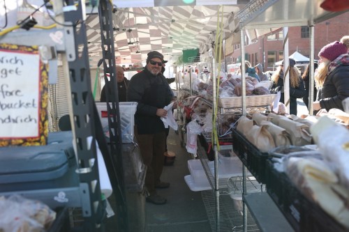 A vendor smiles at a bustling market stall filled with various baked goods and customers browsing.