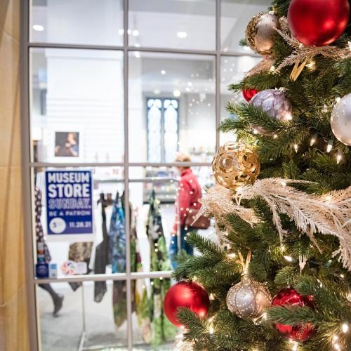 A decorated Christmas tree with ornaments and lights, with a store window displaying a sign for Museum Store Sunday.