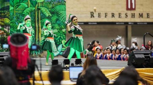 Dancers in vibrant green costumes perform on stage, with a colorful backdrop and an audience watching.