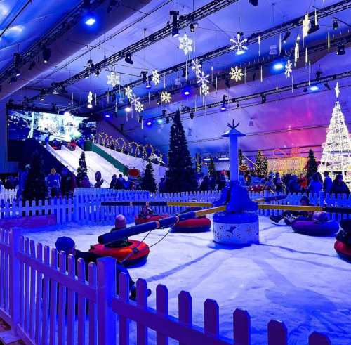 A festive indoor winter scene with snow, a slide, trees, and people enjoying activities under blue lights and snowflakes.