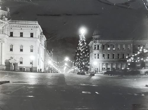 A nighttime street scene decorated with Christmas lights and a large tree, flanked by historic buildings.