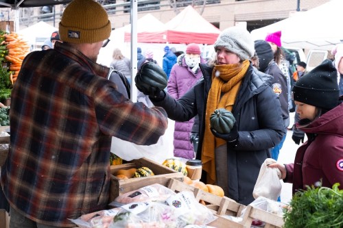 A vendor hands a woman a squash at a bustling outdoor market, with tents and shoppers in the background.