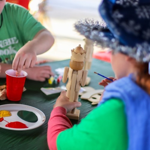 A child paints a wooden figure at a table, with paint palettes and a red cup nearby.