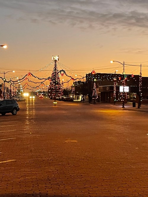A festive street scene at dusk, featuring a decorated Christmas tree and colorful lights along the buildings.