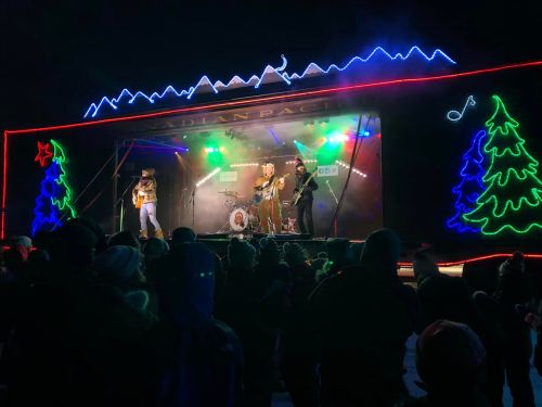 A band performs on a brightly lit stage with festive decorations, while a crowd enjoys the show under a night sky.