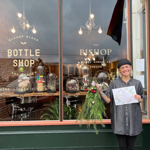 A person stands outside a shop holding a festive arrangement and a certificate, with a window display in the background.