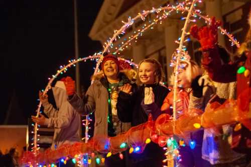 Children smiling and waving on a decorated float with colorful lights during a nighttime parade.