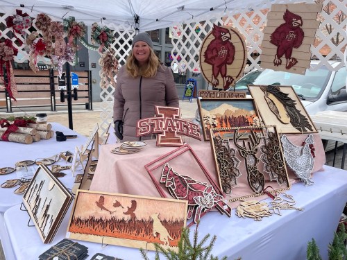 A woman stands behind a table displaying various wooden crafts and decorations at an outdoor market.