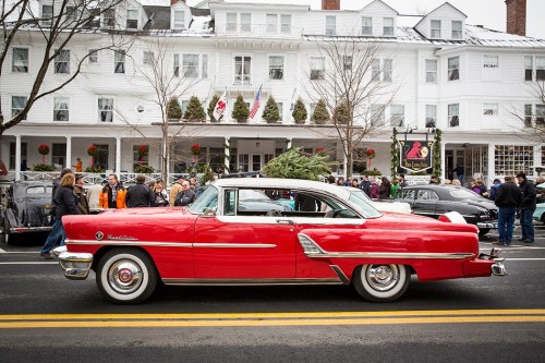 A vintage red car with a Christmas tree on top parked in front of a festive hotel, surrounded by people.