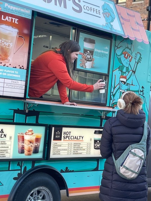 A barista hands a coffee to a customer at a colorful coffee truck. The customer is wearing earmuffs and a backpack.