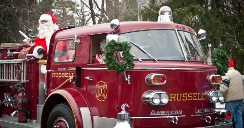 A vintage fire truck decorated for the holidays, with Santa Claus and a passenger wearing festive attire.