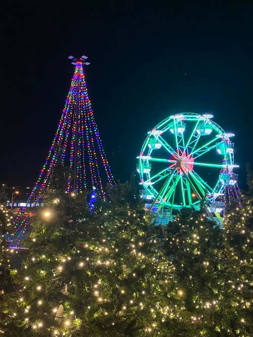 A brightly lit Christmas tree and a colorful ferris wheel at night, surrounded by festive lights.