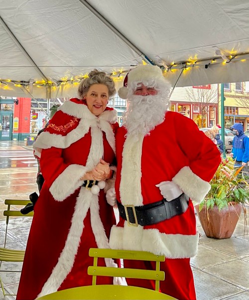 A man and woman dressed as Santa and Mrs. Claus pose together under a festive tent.